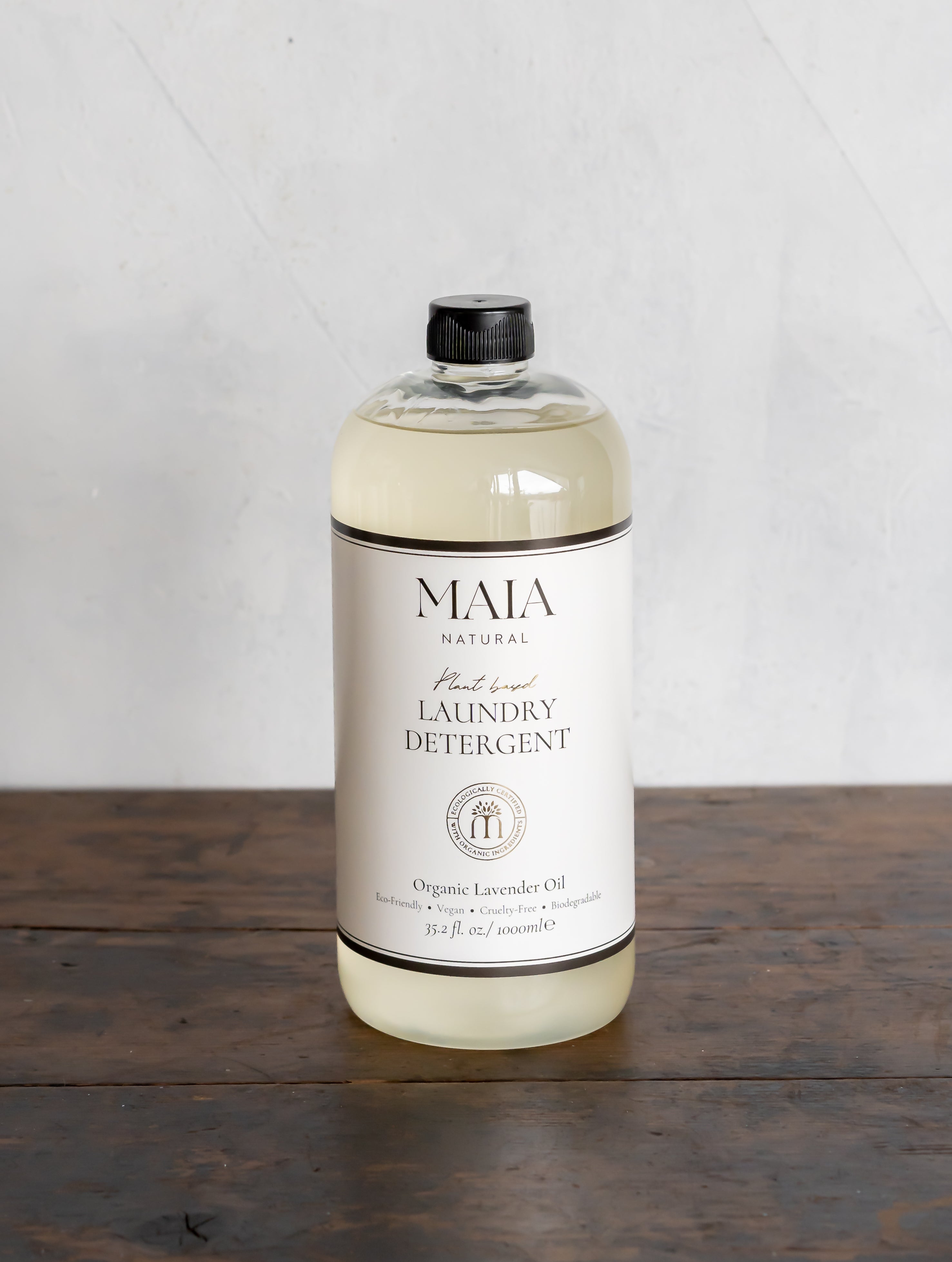 A clear bottle of Maia Natural Laundry Detergent with a black cap, standing on a wooden surface with a white label that includes the product name and description.