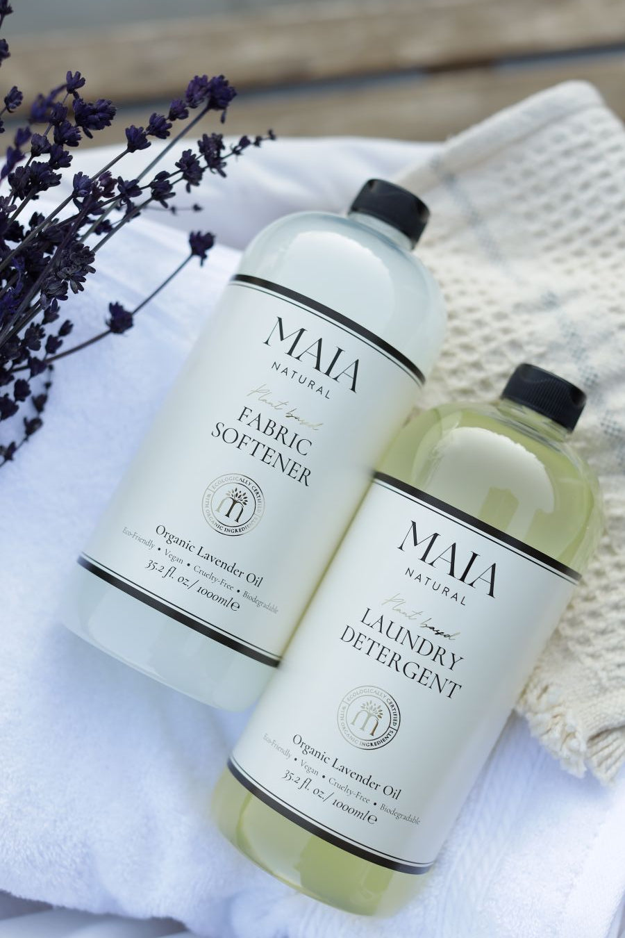 Two bottles of MAIA natural laundry softener and detener on a white linen fabric backdrop with some purple flowers.